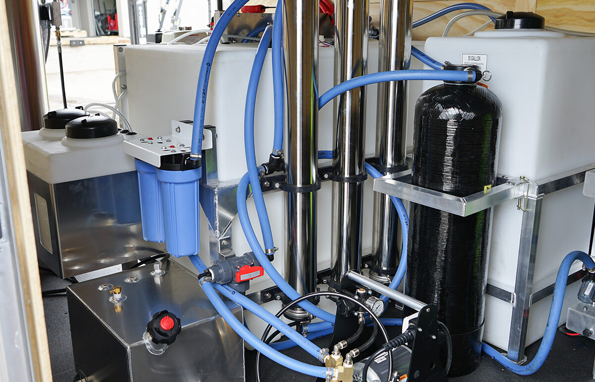 Industrial water filtration system: vertical stainless filter housings, blue hoses, white reservoir, and a pump in a workshop setup.